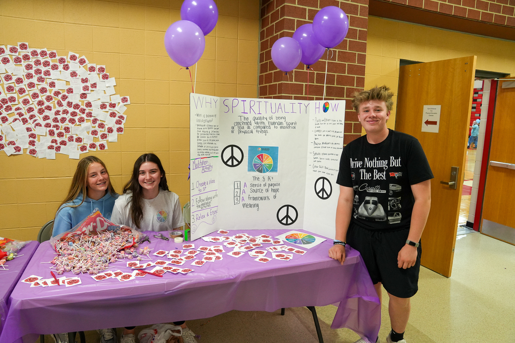 Two students sit behind a table with a display board about spirituality, surrounded by purple decorations and candy for an activity station.