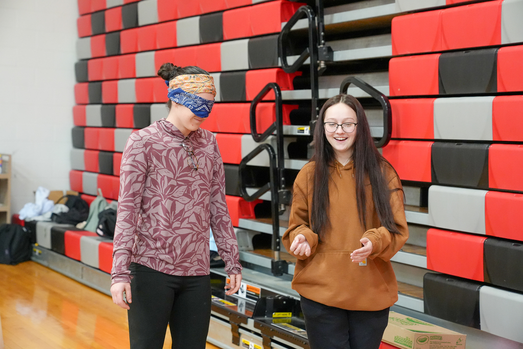 Two students walk side by side in the gym, one blindfolded, as part of a trust-based activity.