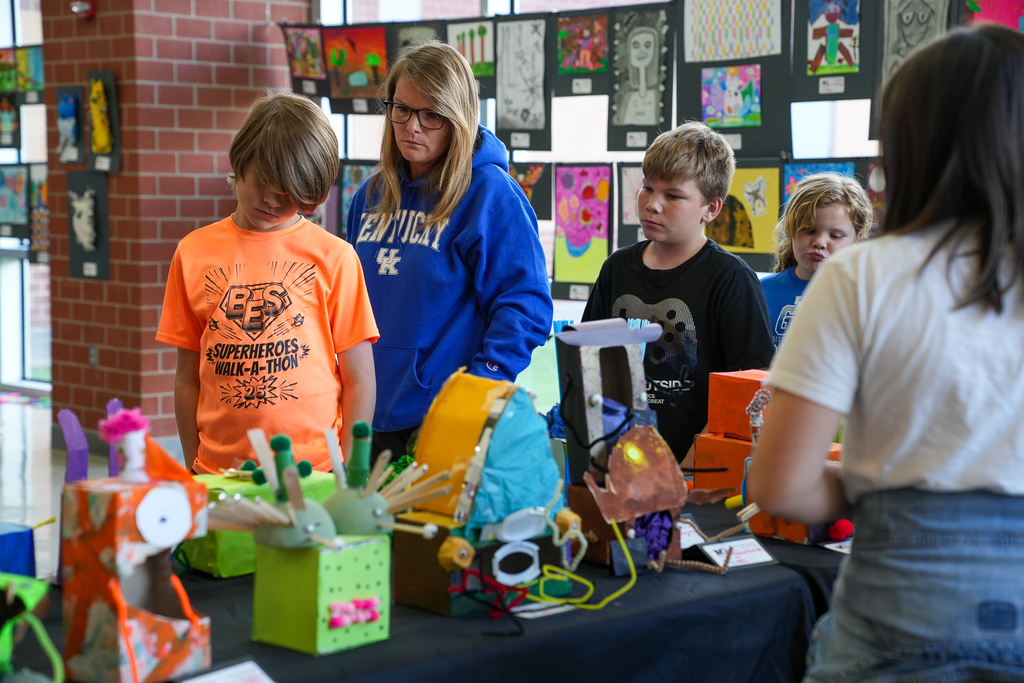 A group of students and an adult look at colorful, handmade art projects displayed on a table, including creative, three-dimensional pieces.