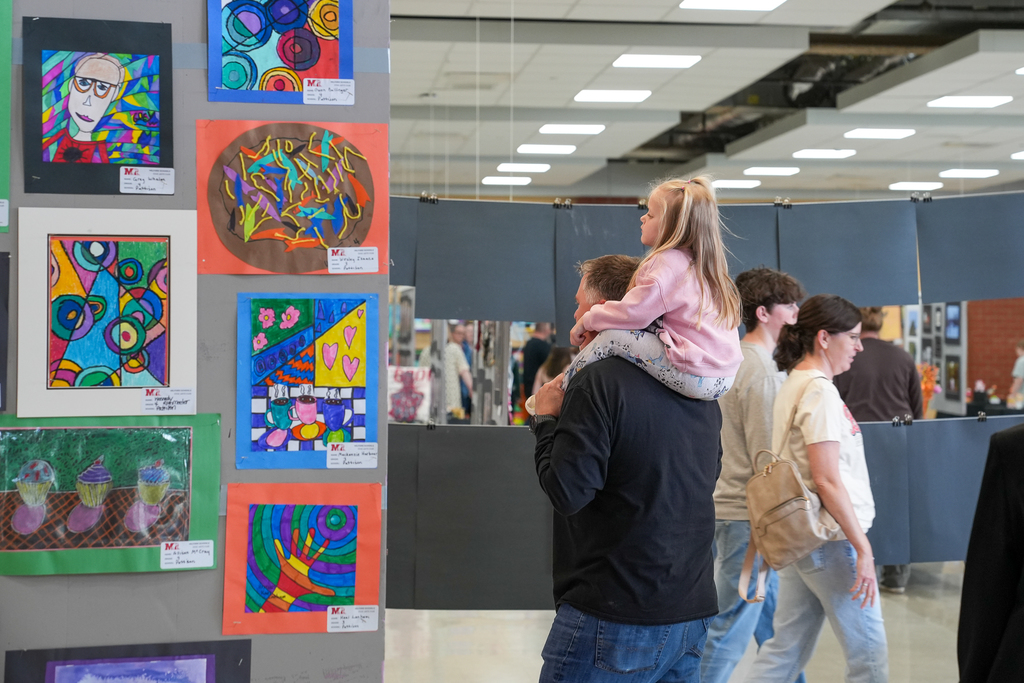 A man holds a young child on his shoulders as they look at a display of vibrant student artwork in a school setting, while other visitors walk through the exhibit.