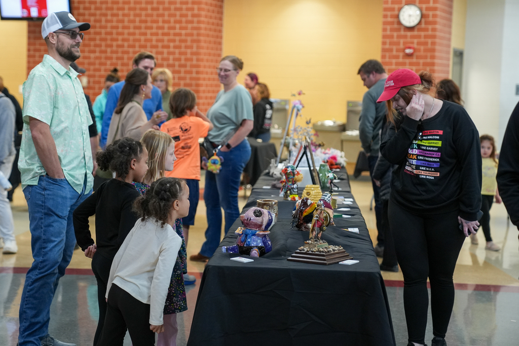 Students and adults gather around a table displaying small sculptures and creative projects, examining the pieces closely in a school art exhibit.