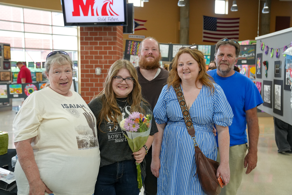 A group of five people stands together smiling for a photo at a school art show, with student artwork displayed behind them.