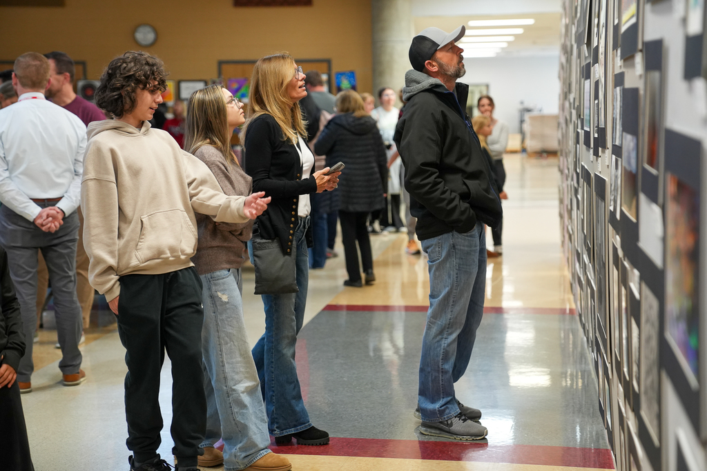 Three people stand side by side in a school hallway, closely examining a large display of framed student artwork.