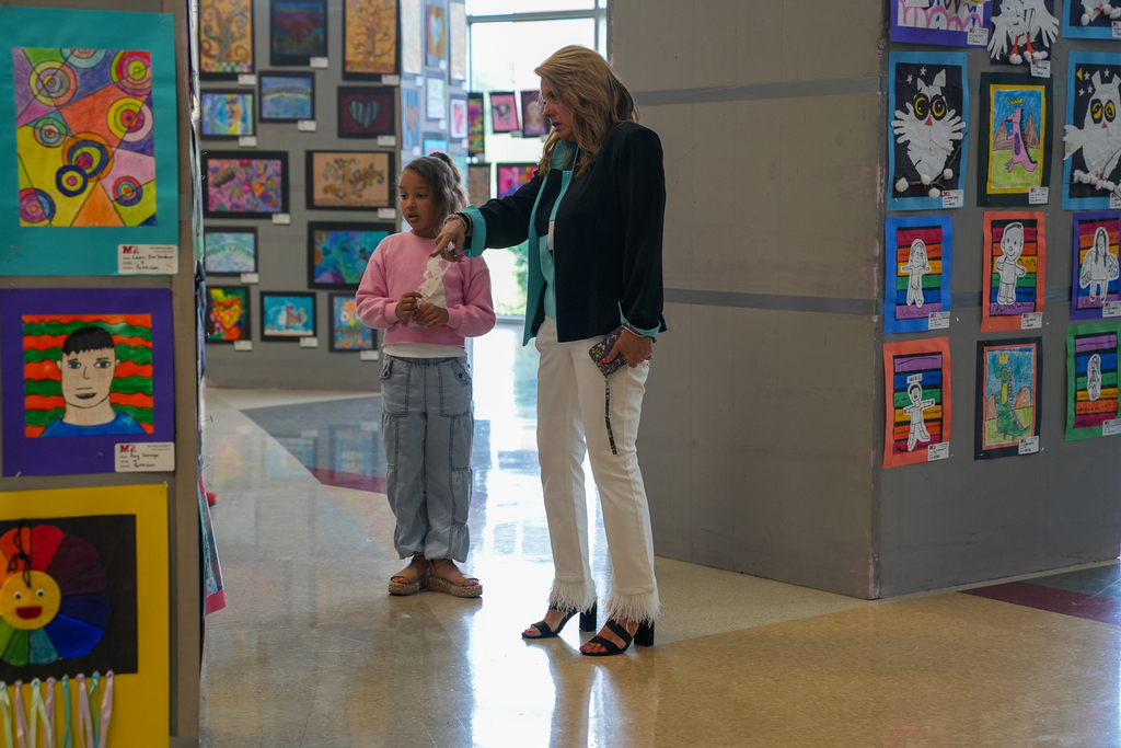 An adult and a young student stand together in a school hallway, pointing and discussing student artwork displayed on large panels.