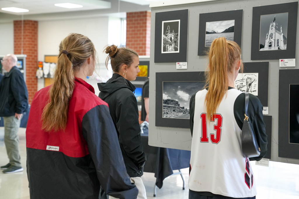 Three students stand in front of a display of black-and-white photography, studying the images mounted on a panel at a school art show.