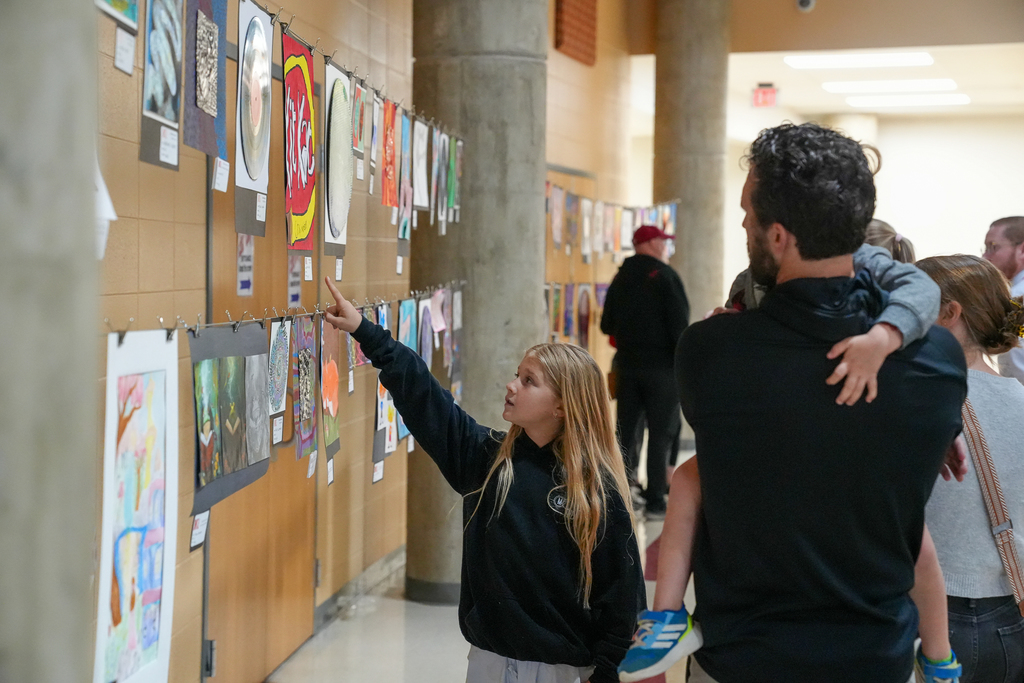 A young student points at artwork displayed along a hallway wall while standing next to an adult holding a child, both observing the exhibit.