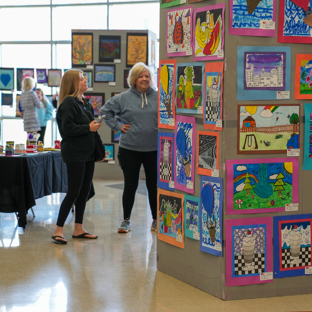Two adults stand in a school hallway viewing a display wall filled with colorful student artwork, including drawings and paintings mounted on bright paper backgrounds.