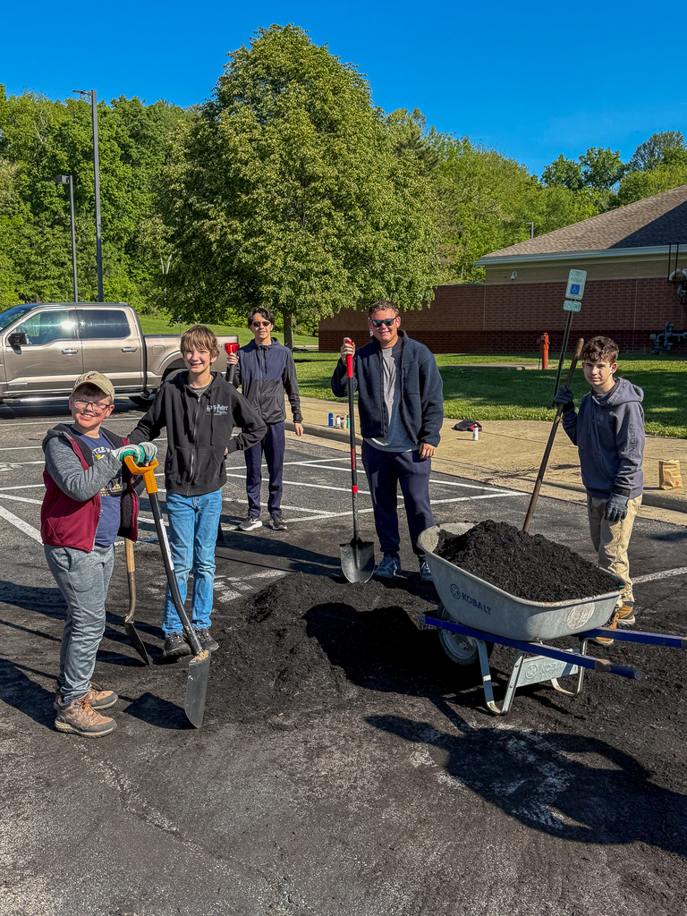 A group of students and an adult stand in a parking lot with shovels and a wheelbarrow full of mulch, smiling while working on a landscaping project.