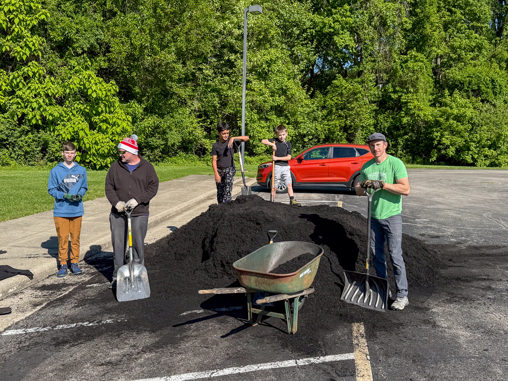 A group of volunteers, including adults and students, stand around a large pile of mulch with shovels and a wheelbarrow in a parking lot near trees.
