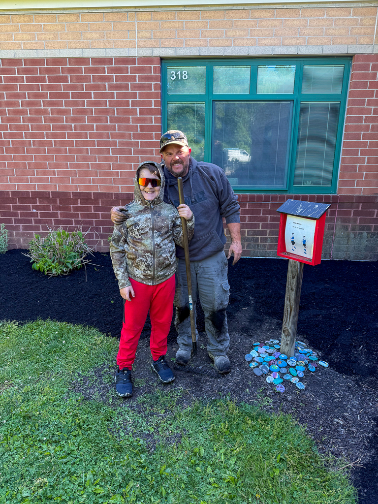 An adult and a child stand together smiling near a freshly mulched garden bed, holding a shovel beside a small post with painted rocks at its base.