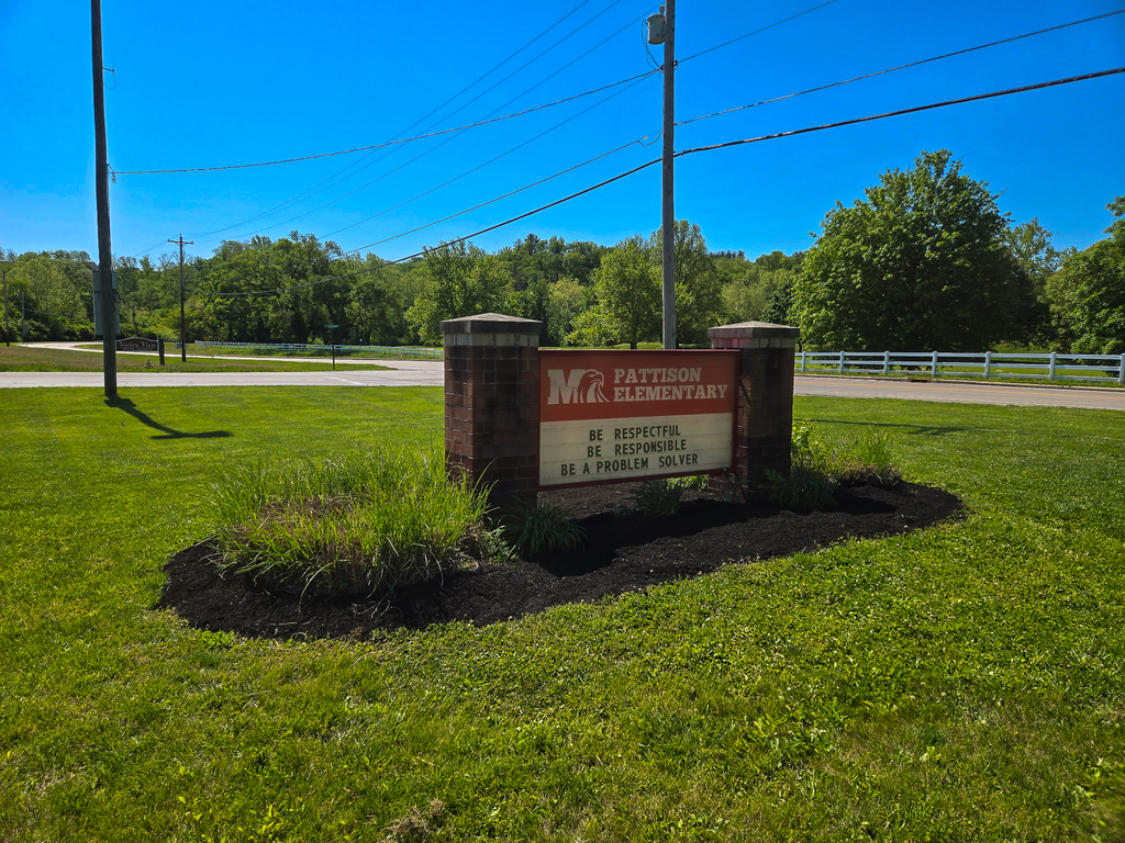 A brick Pattison Elementary sign sits along a roadside, surrounded by freshly spread mulch and trimmed landscaping under a bright blue sky.