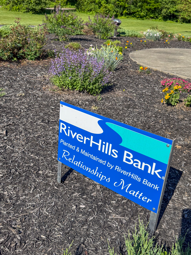 A small sign in a mulched flower bed reads “RiverHills Bank – Planted & Maintained by RiverHills Bank, Relationships Matter,” with blooming plants and greenery behind it.