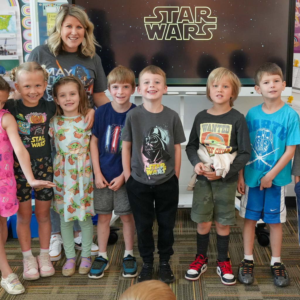A teacher stands smiling behind a group of elementary students lined up in a classroom, all facing the camera. Several students are wearing Star Wars-themed clothing, including shirts featuring characters like Darth Vader and Grogu. Behind them, a large screen displays the “Star Wars” logo against a starry background. The classroom walls show colorful learning materials, and the students appear cheerful and excited.