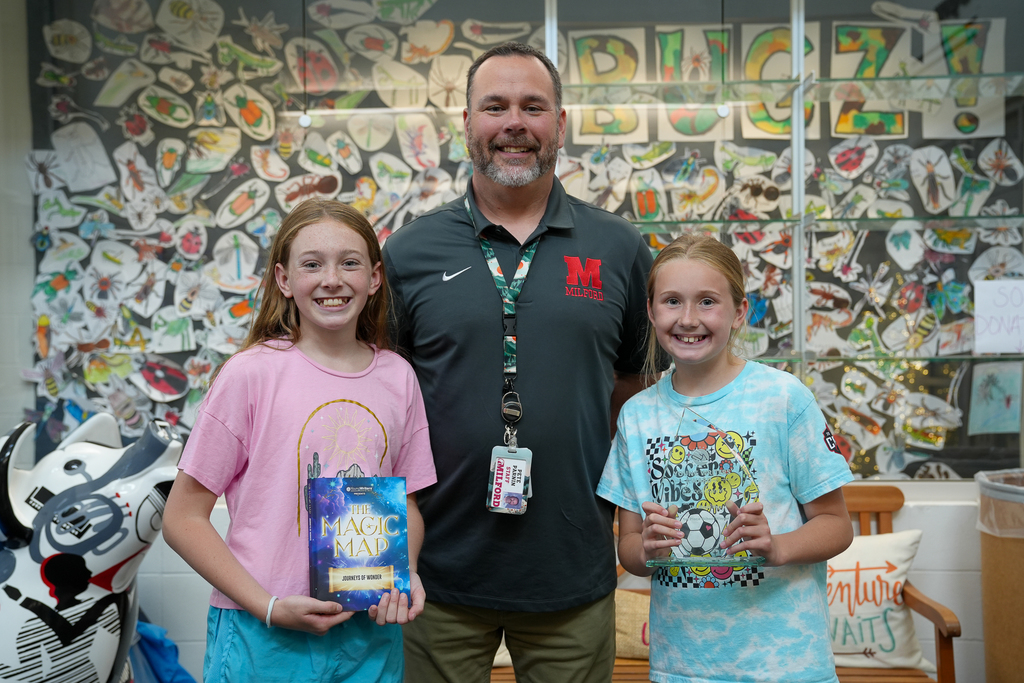 A teacher stands smiling between two elementary-aged students in a school hallway or classroom display area. The student on the left holds a book titled The Magic Map: Journeys of Wonder, while the student on the right holds a clear award plaque. All three face the camera and smile. Behind them is a colorful wall filled with student artwork featuring insects and drawings, along with the word “BUGZ” displayed across the top.