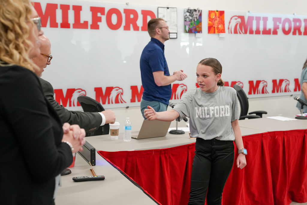 A student wearing a "Milford" shirt focuses while fist bumping a school administrator during a recognition at a Board of Education meeting, with a “Milford” sign visible in the background.