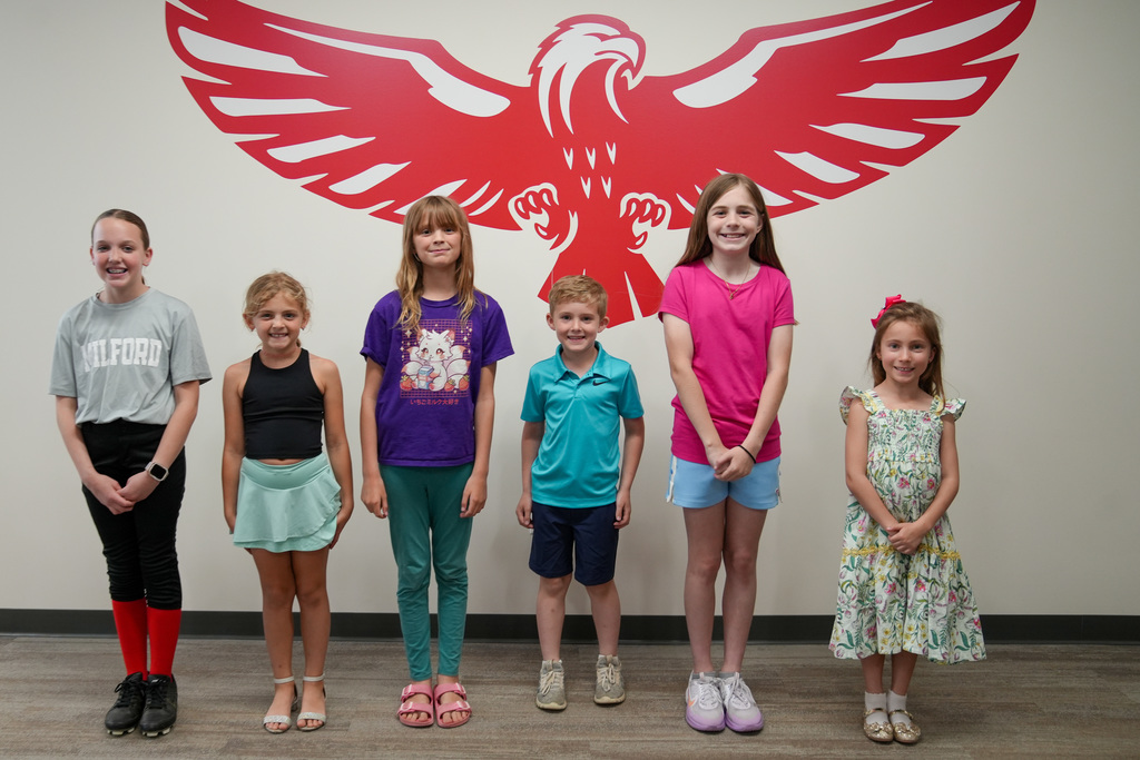 Six elementary-aged students stand in a line in front of a large red eagle graphic on a wall. The students are smiling and dressed casually, posing for a recognition photo.