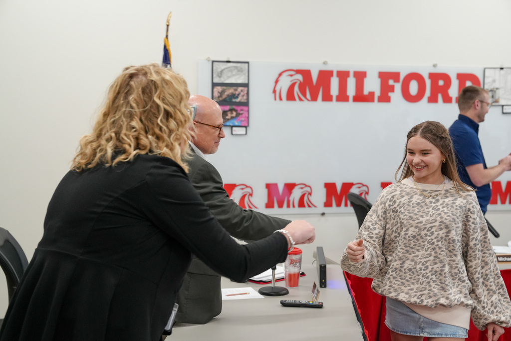 A student wearing a patterned sweatshirt smiles while fist bumping a school administrator during a recognition at a Board of Education meeting, with a “Milford” sign visible in the background.