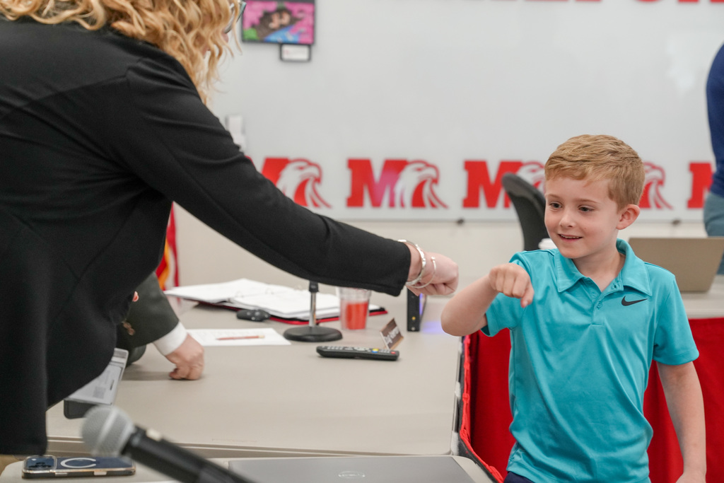 A young student in a teal shirt fist bumps a school administrator across a table during a recognition moment at a Board of Education meeting.