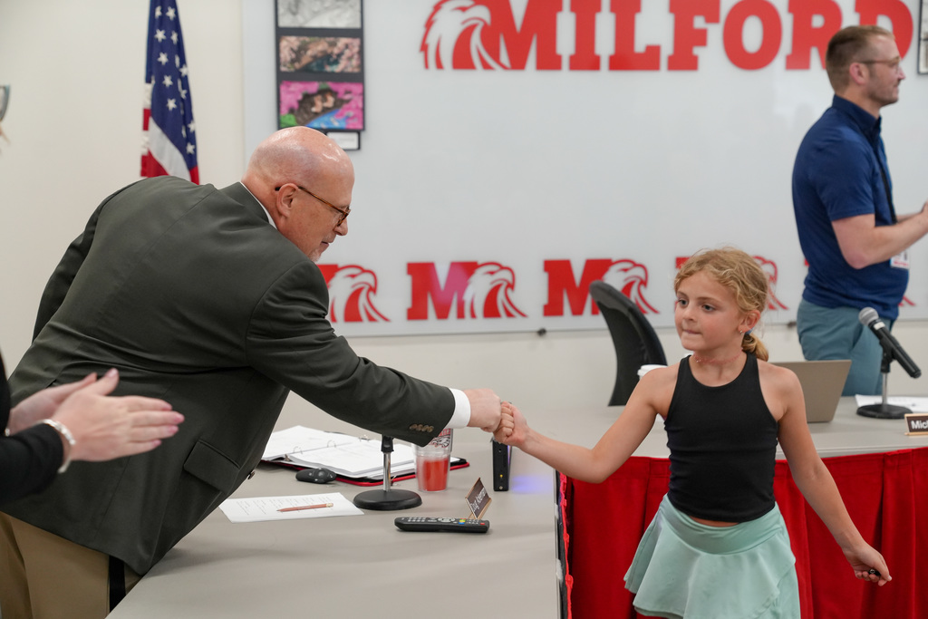 A young student in a black tank smiles while fist bumping a school administrator at the front of a meeting room during a recognition ceremony.