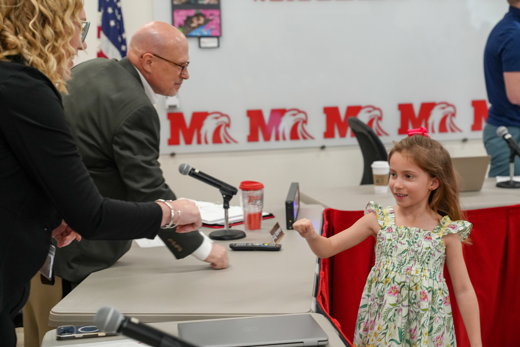 A young student in a floral dress smiles while fist bumping a school administrator at the front of a meeting room during a recognition ceremony.