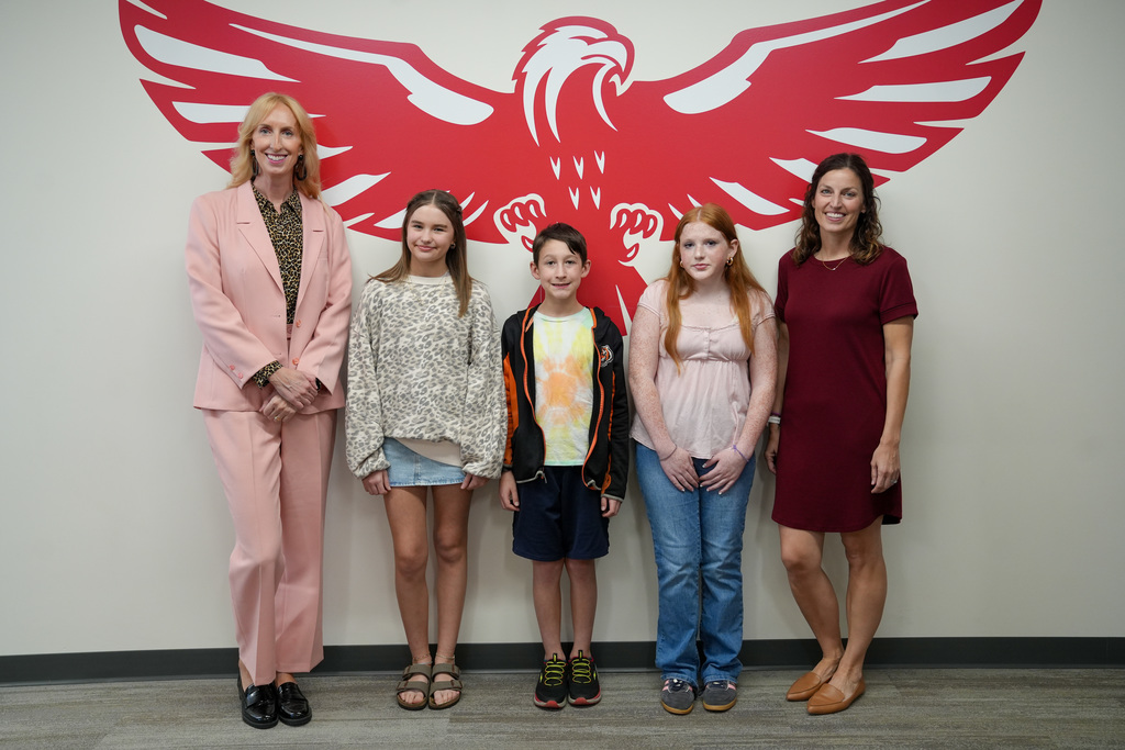 Two school administrators stand with three students in front of a large red eagle wall graphic. The group poses for a photo, smiling, as part of a recognition at a Board of Education meeting.