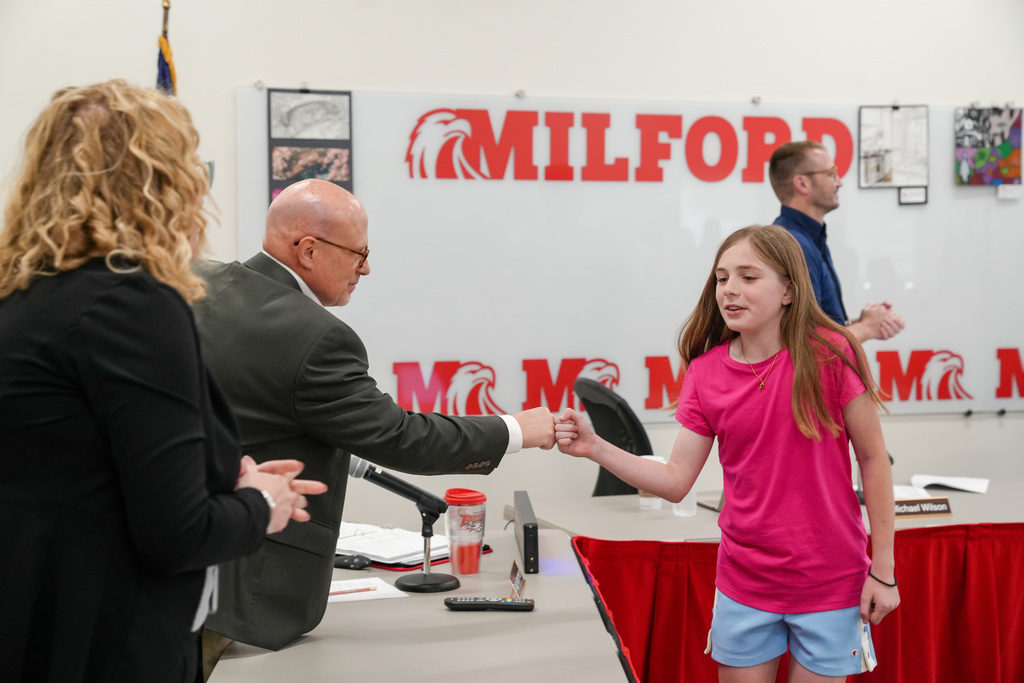 A student in a pink shirt fist bumps a school administrator at a table during a Board of Education meeting. Other staff members stand nearby, and a “Milford” sign is visible on the wall behind them.