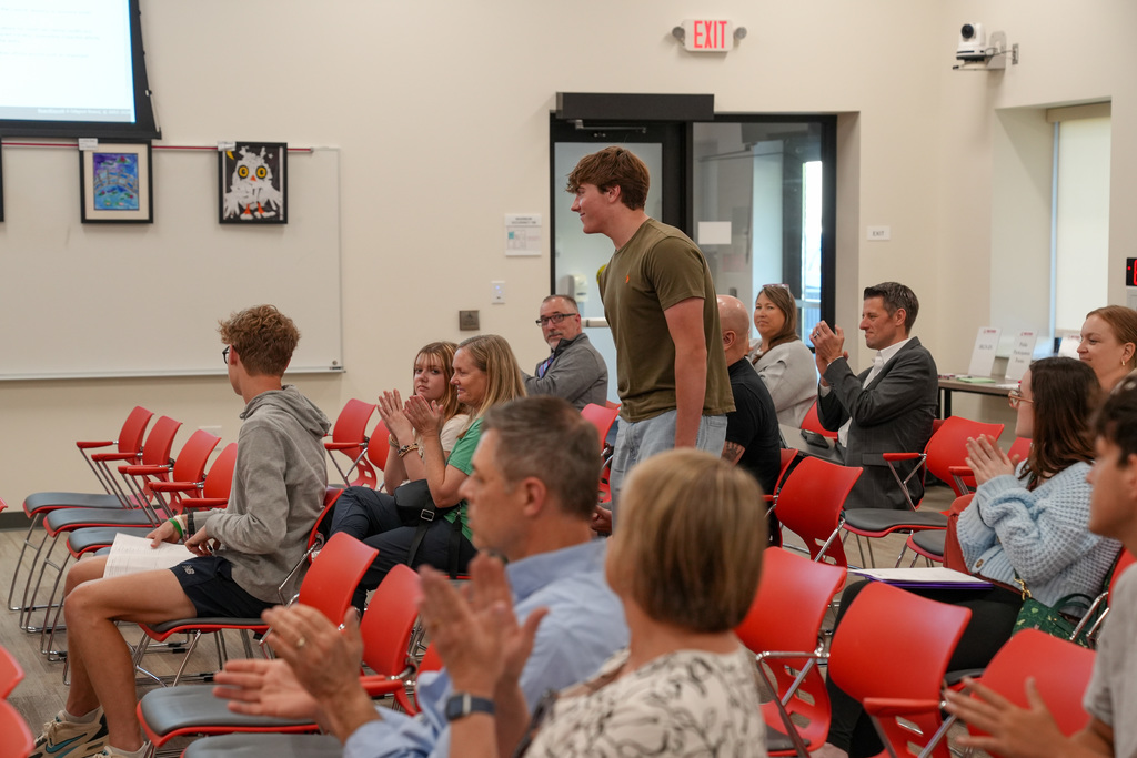 A high school student stands in a meeting room filled with red chairs as attendees applaud. Several adults and students are seated around him, clapping in recognition during a Board of Education meeting.A high school student stands in a meeting room filled with red chairs as attendees applaud. Several adults and students are seated around him, clapping in recognition during a Board of Education meeting.