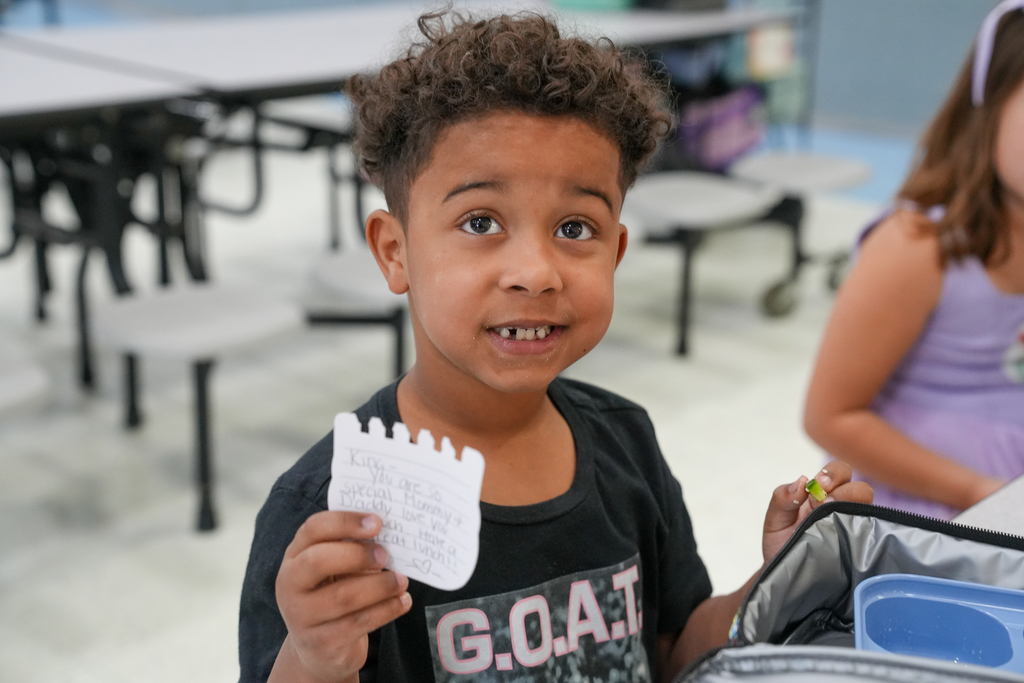 A smiling kindergarten student holds up a handwritten note while sitting in the cafeteria with classmates during lunch.