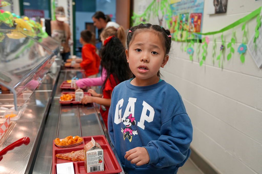 A young student wearing a blue GAP sweatshirt stands in the cafeteria line with a lunch tray containing pizza, tater tots, and milk while classmates wait behind her.