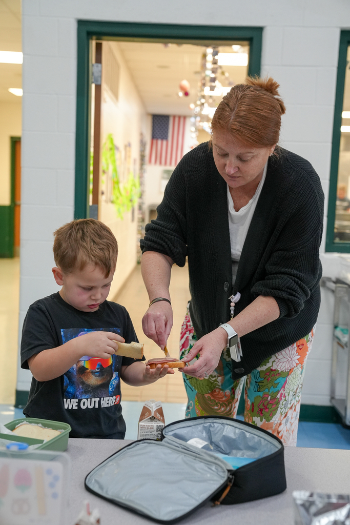 Principal Greb assists a kindergarten student spreading mustard onto bread at a cafeteria table while the student prepares his lunch.