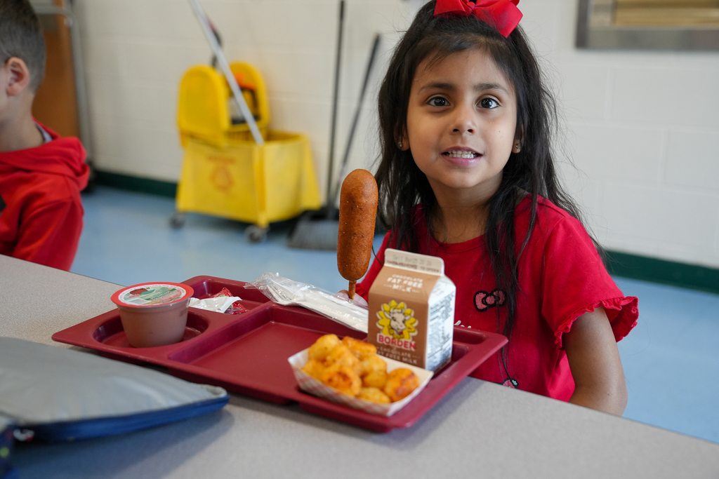A smiling kindergarten student wearing a red shirt and bow sits at a cafeteria table with her lunch tray, holding up a corndog for the camera.