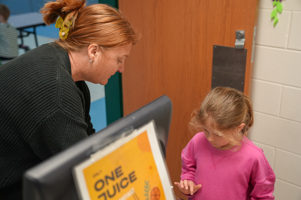Principal Greb  kneels beside a kindergarten student in the cafeteria while helping her through the lunch process. A sign for juice options is visible in the foreground.