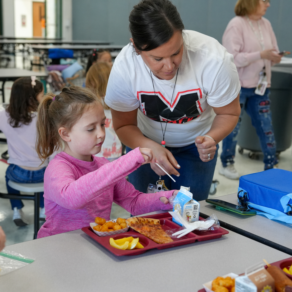 Ms. Cowell helps a kindergarten student open and organize items on her lunch tray while they sit together in the cafeteria during the class lunch practice.