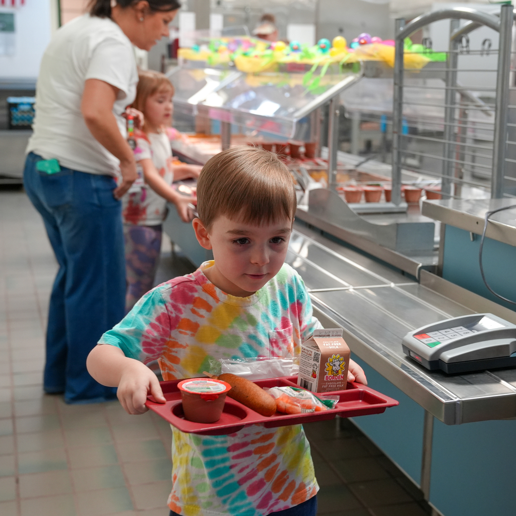 A kindergarten student wearing a colorful tie-dye shirt carefully carries a red lunch tray with milk, vegetables, and a corndog through the cafeteria line while staff assist students in the background.