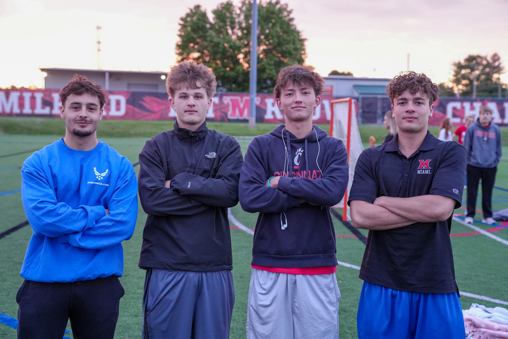 Four male Milford seniors stand on the turf field with their arms crossed during Senior Sunrise. The students wear casual athletic clothing and college apparel, including Cincinnati and Miami University gear, while posing in front of the Milford athletics signage at sunrise.