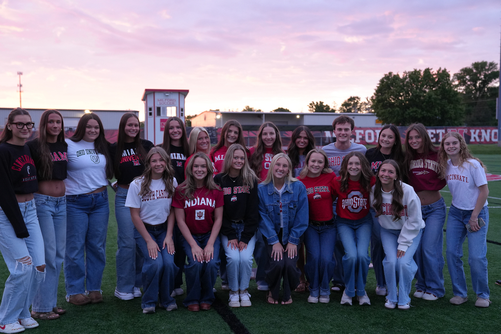 A large group of Milford High School seniors pose together on the turf field during Senior Sunrise at sunrise. Students wear college and university apparel representing schools such as Ohio State, Miami University, Cincinnati, Indiana, and South Carolina while smiling for a group photo under a pink and purple morning sky.