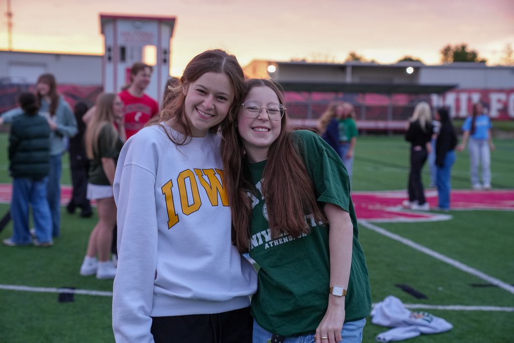 Two Milford seniors smile with their arms around each other during Senior Sunrise on the turf field. One student wears an Iowa sweatshirt while the other wears an Ohio University shirt, with other students blurred in the background during the early morning event.
