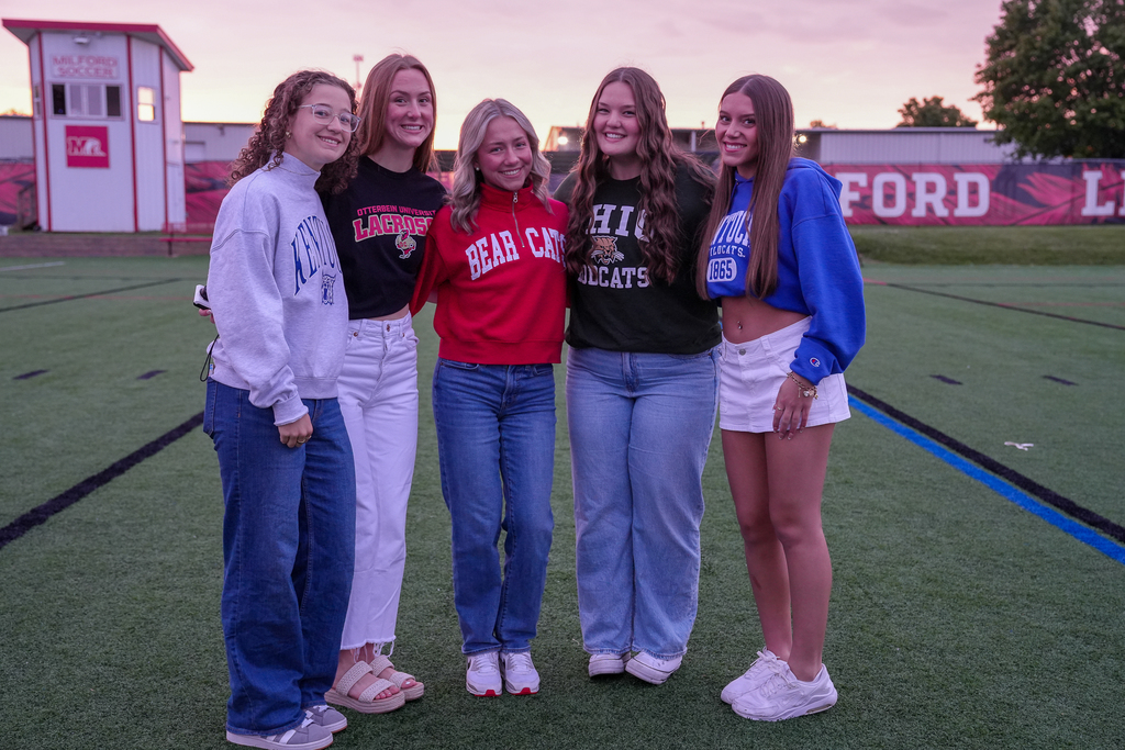 Five female Milford seniors smile together on the turf field during Senior Sunrise while wearing college and university apparel representing schools including Kentucky, Otterbein, Ohio, and Cincinnati. The pink sunrise sky glows behind them.