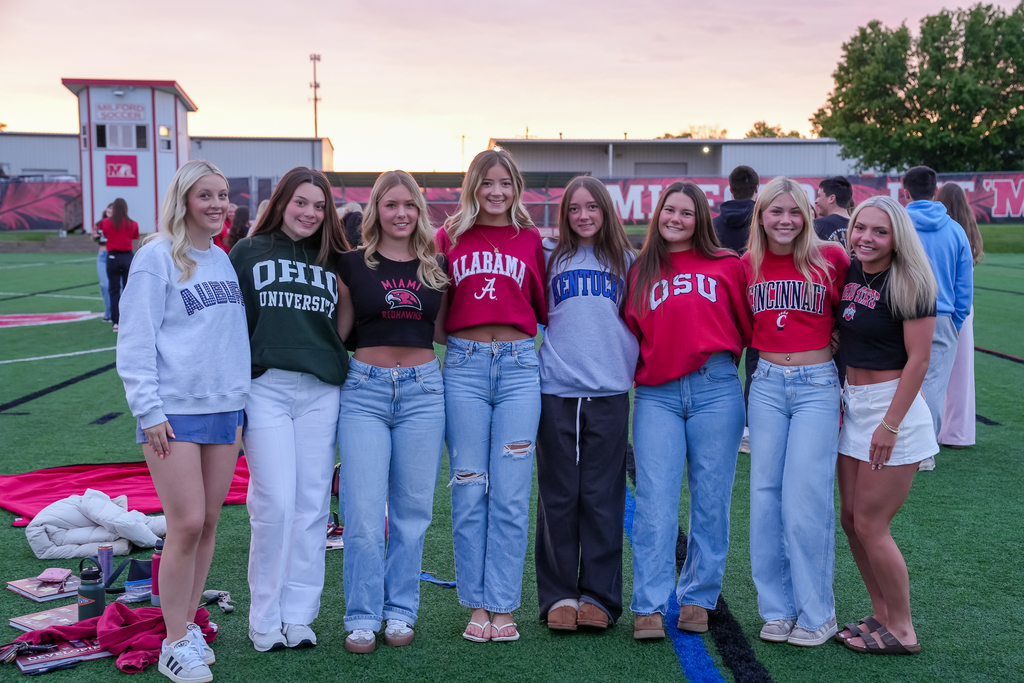 Seven female Milford seniors pose together on the turf field during Senior Sunrise while wearing college apparel from schools including Auburn, Ohio University, Alabama, Kentucky, Ohio State, Cincinnati, and Ohio State. Blankets and personal items are scattered on the turf behind them as the sun rises.