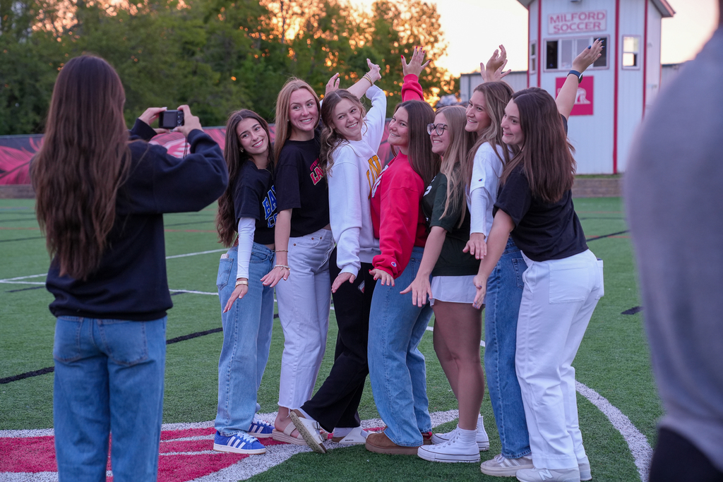 A group of Milford seniors pose together for a photo during Senior Sunrise while another student takes their picture. The students smile and hold playful poses on the turf field as the sun rises behind the trees.