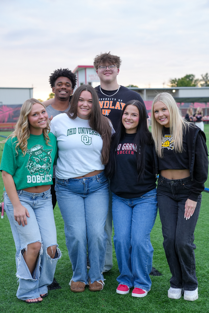 A group of Milford seniors wearing college apparel from schools including Ohio University, Ohio Northern, Rio Grande, and Northern Kentucky University smile together for a group photo during Senior Sunrise on the turf field.