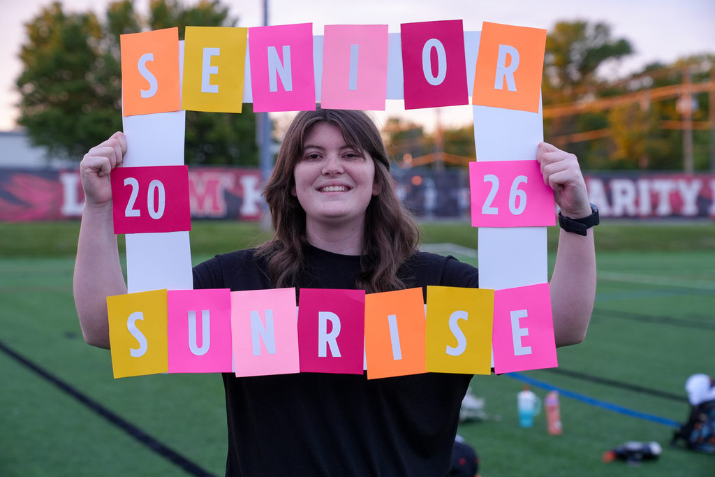 A Milford senior smiles while holding a colorful handmade frame that reads “Senior Sunrise 2026” during the early morning event on the turf field.