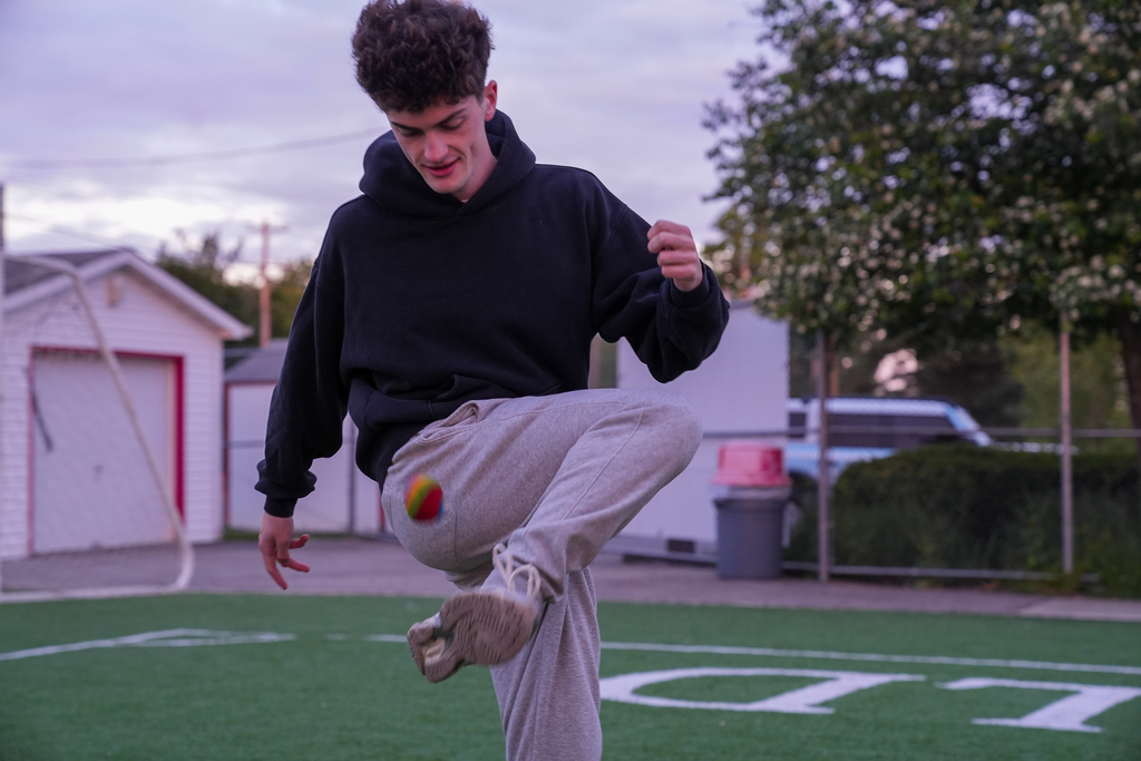 A Milford senior plays with a colorful ball on the turf field during Senior Sunrise while wearing a black hoodie and gray sweatpants. The early morning sky and trees are visible in the background.