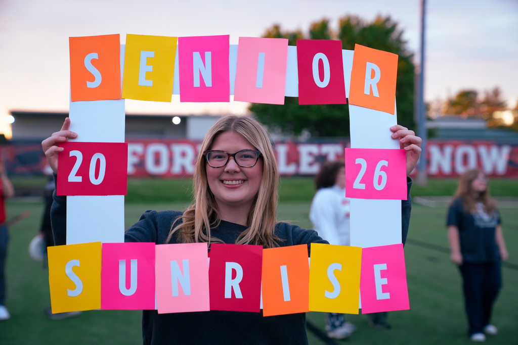 A Milford senior wearing glasses smiles while holding a colorful “Senior Sunrise 2026” frame on the turf field during the sunrise celebration.