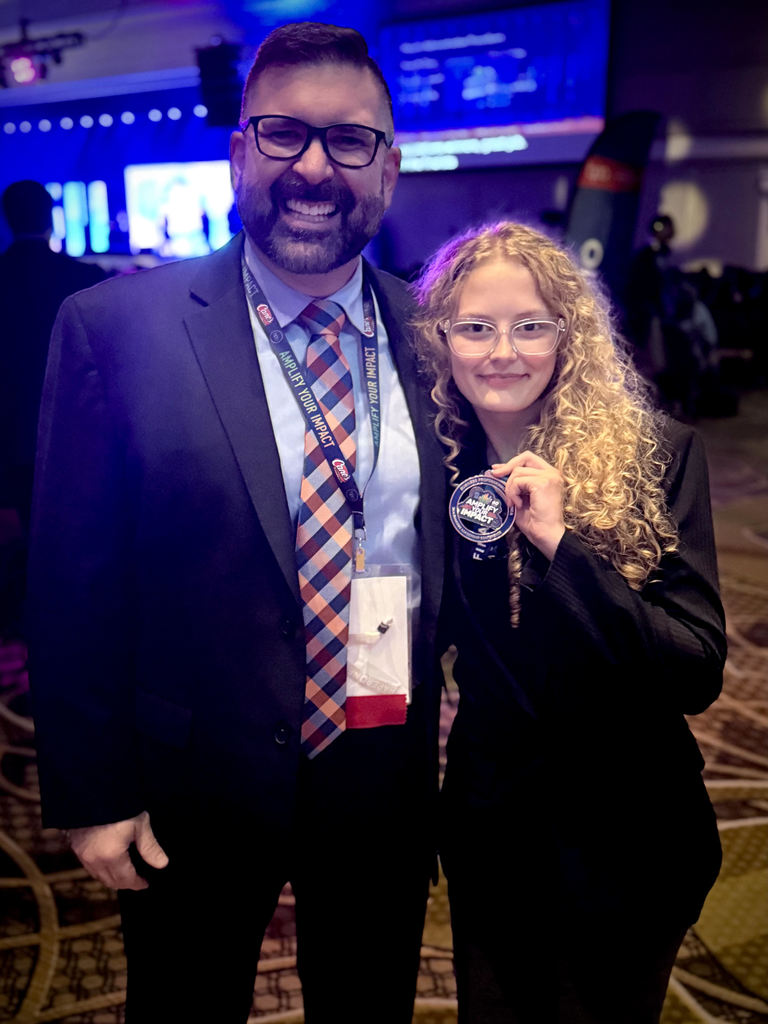 A student and staff member smile together at an event venue with blue and purple lighting in the background. The student, wearing a black outfit and glasses, holds up a medal while standing beside the staff member in a suit and patterned tie.