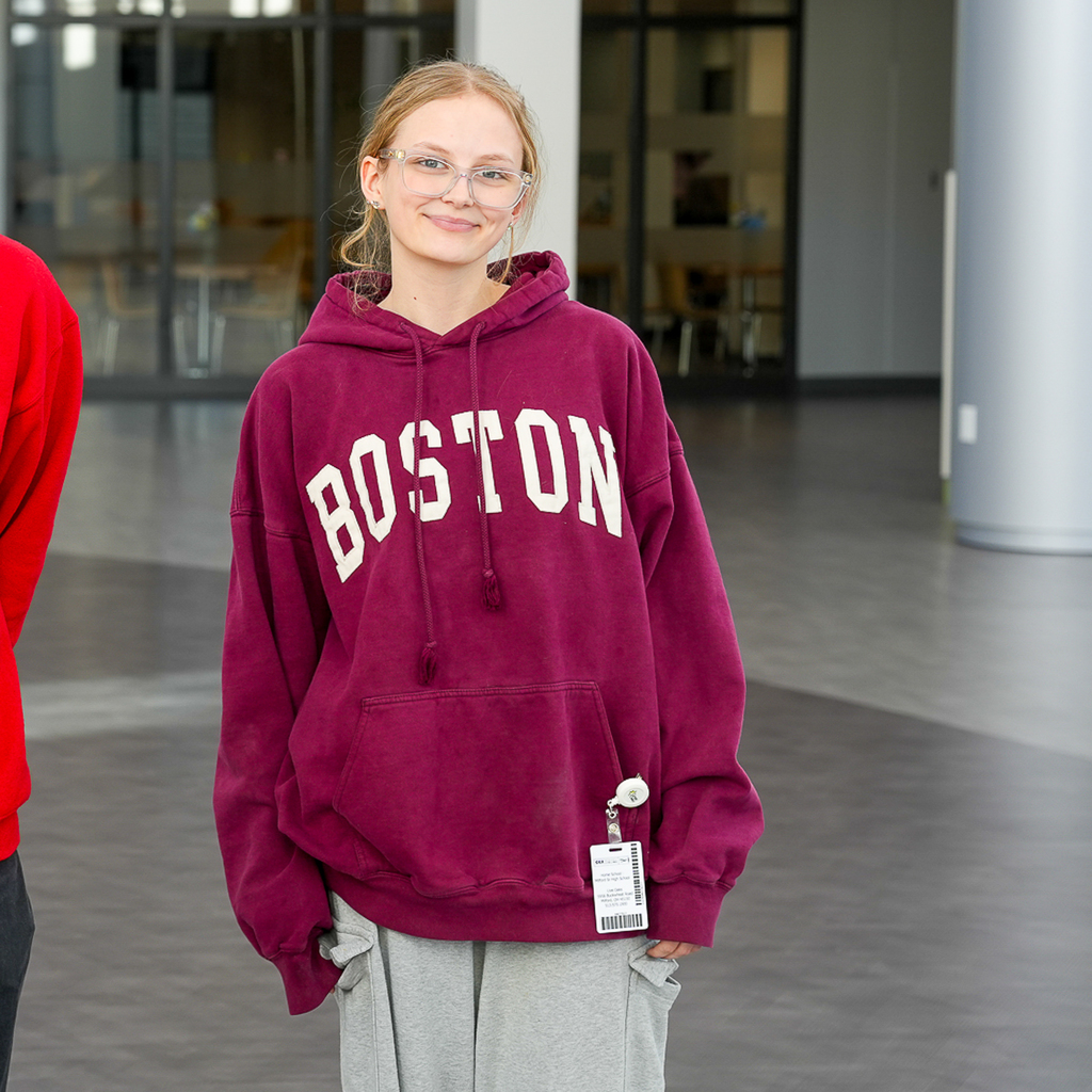 A student wearing glasses stands in a bright open hallway wearing a maroon “Boston” hoodie and gray sweatpants. A school ID badge is clipped near the pocket of the sweatshirt.
