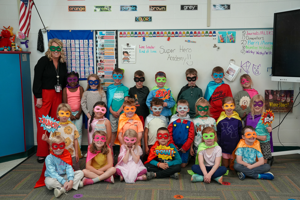 A class of kindergarten students dressed as superheroes pose together in front of a comic book-themed backdrop while wearing colorful masks and capes.
