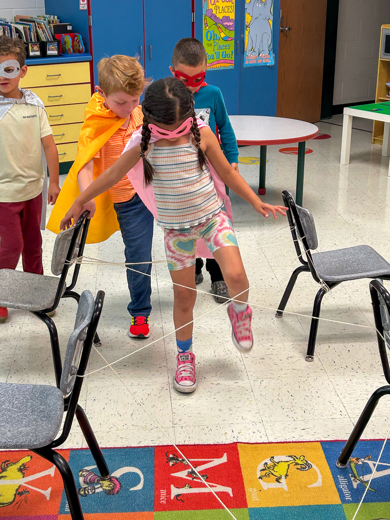 Kindergarten students dressed in colorful superhero capes and masks carefully work together to navigate through a classroom obstacle course made of string and chairs.