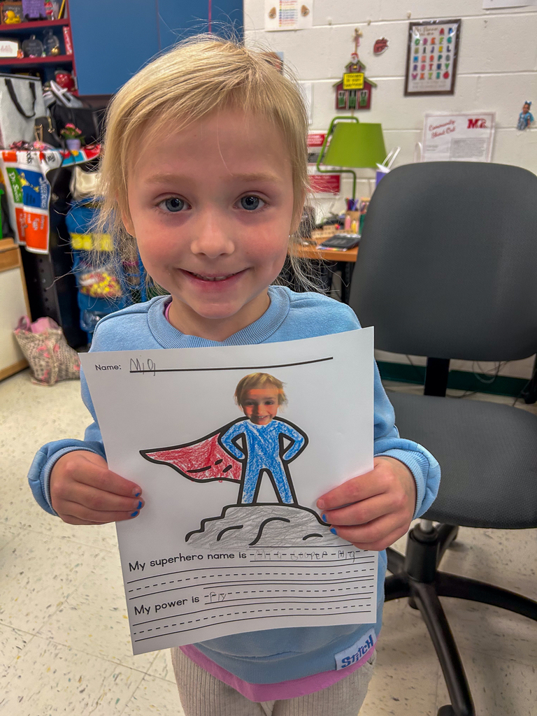 A kindergarten student smiles while holding a “My Superhero” worksheet featuring a drawing of themselves as a superhero with a red cape and blue suit. The student’s superhero power is listed as “fly.”