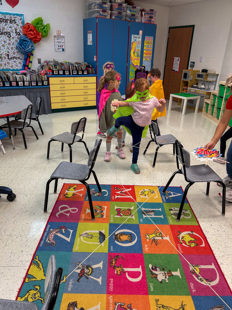 Several kindergarten students in superhero costumes make their way through a classroom “laser maze” obstacle course during Superhero Academy activities.